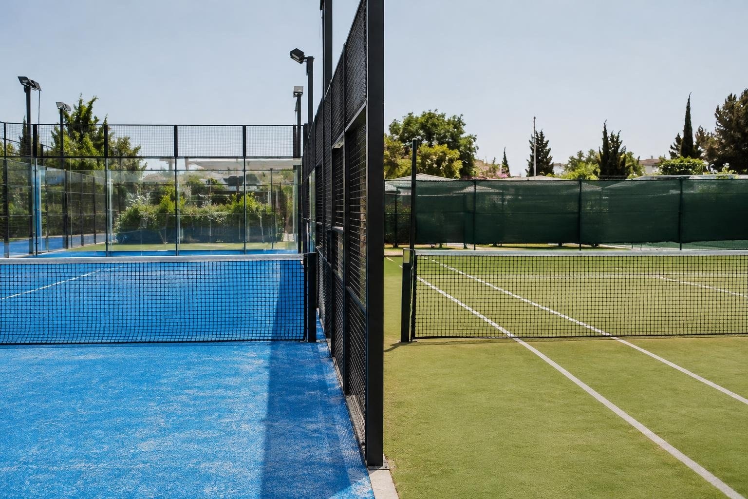 Padel vs Tennis: What Is the Difference? Two empty racket courts sit side by side, one with a blue surface and glass walls and the other with a green surface, surrounded by fences, trees, and light poles.